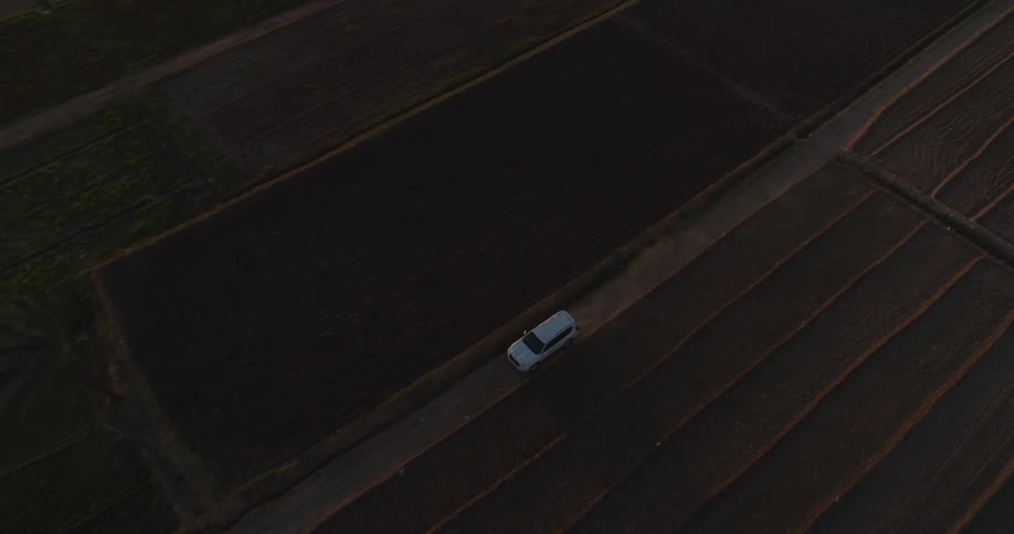 An aerial view shows a white car driving along a dirt road through plowed agricultural fields during a golden sunset