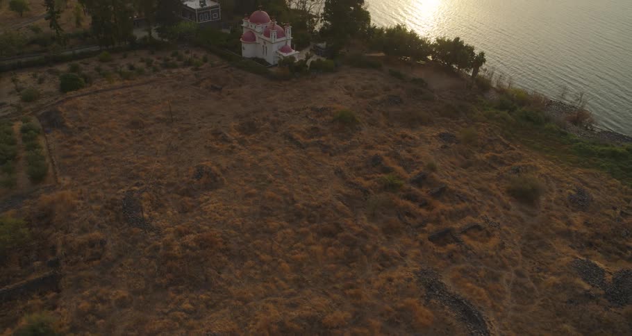 Aerial shot reveals the Greek Orthodox Church of the Twelve Apostles next to the ruins of Capernaum on the Sea of Galilee during a golden sunset