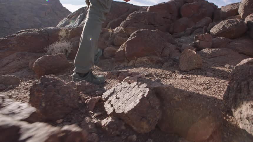 Slow motion low angle shot of a hiker's boots climbing over rugged rocks on a sunny mountain trail