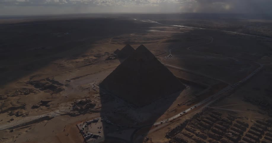 A dramatic high-angle aerial shot shows the Great Pyramids of Giza silhouetted against the bright sun