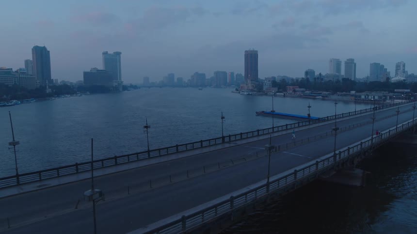 An aerial view shows a lone jogger on an empty bridge crossing a wide river into a city skyline at dawn