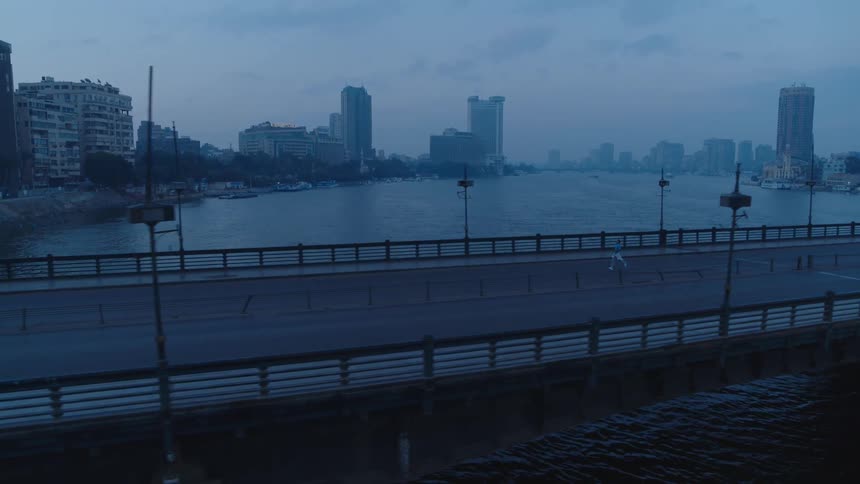 An atmospheric aerial shot follows people jogging across a city bridge over a wide river at dawn