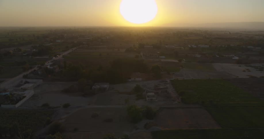 Aerial pullback shot reveals a vast rural landscape of fields and villages under the hazy golden light of a beautiful sunset