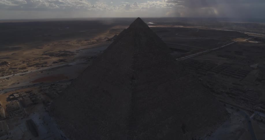 A dramatic aerial view slowly pulls back from the silhouetted Pyramids of Giza as sun rays break through the clouds over the Egyptian desert