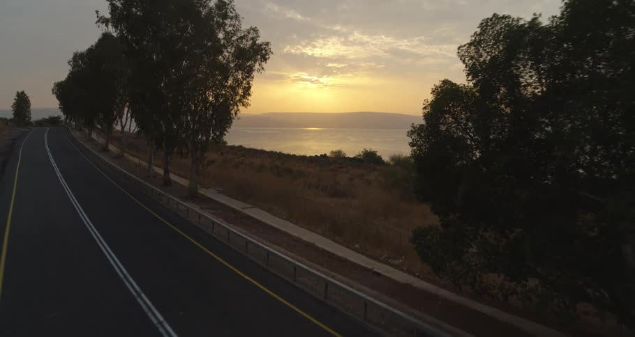 Scenic aerial drone footage flies over a tree-lined road revealing the Sea of Galilee at sunrise