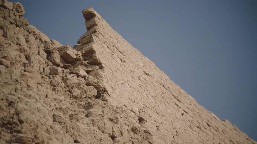 Slow tilt up a crumbling ancient adobe wall revealing its weathered texture against a clear blue sky
