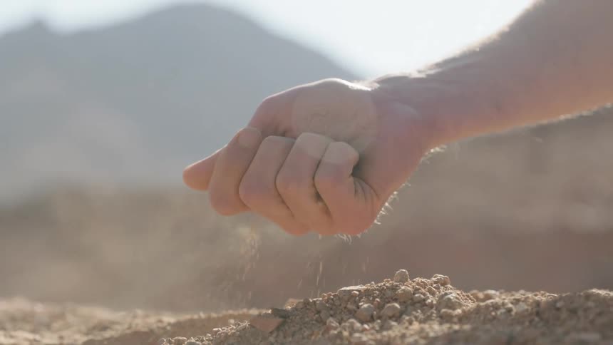 A man's hand slowly lets dry desert soil sift through his fingers in the bright sunlight, filmed in slow motion