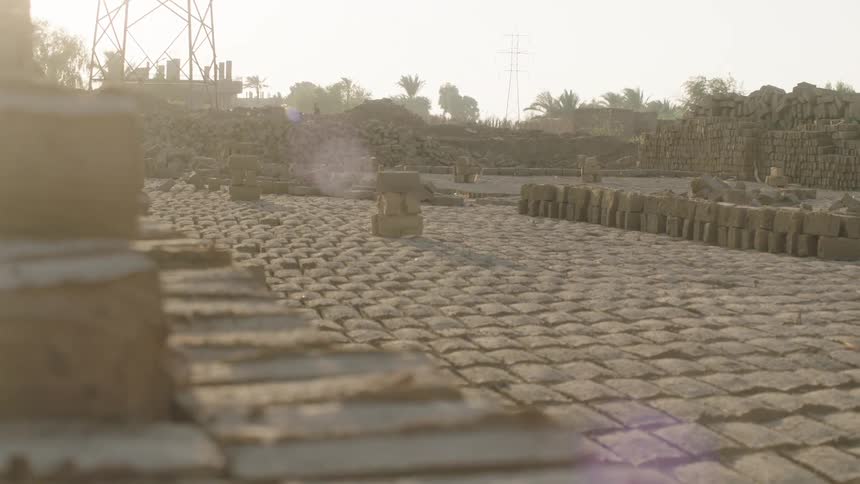 Sunlight flares over countless handmade mud bricks drying in orderly rows at an outdoor brickyard