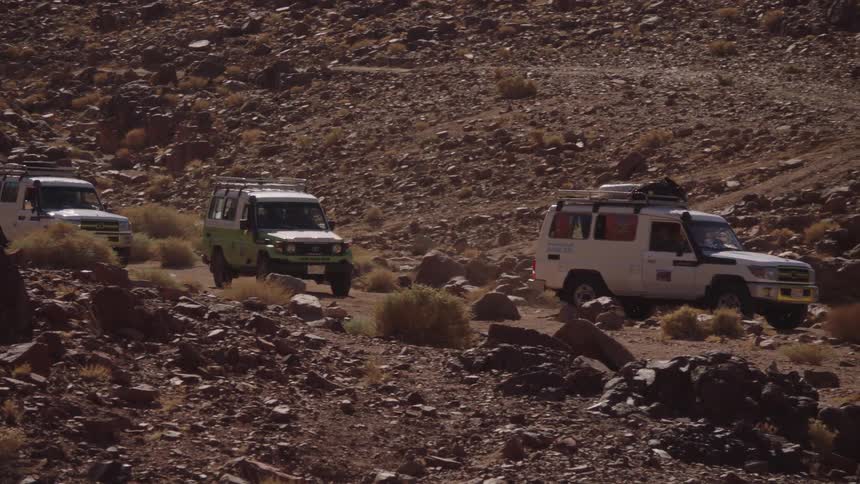 A convoy of rugged 4x4 vehicles slowly navigates a winding, rocky dirt road through a remote desert canyon