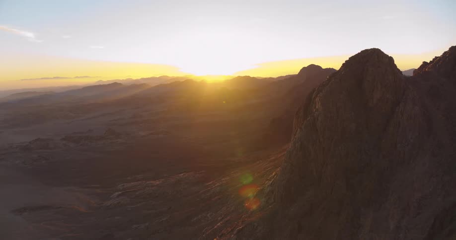 An epic aerial shot slowly pans right across a vast desert mountain range as the sun rises on the horizon