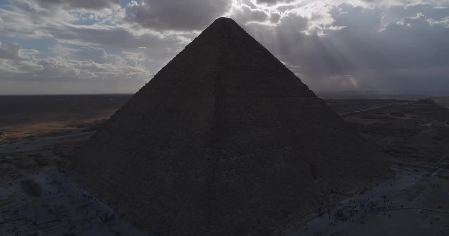 A dramatic aerial view shows the Great Pyramid of Giza silhouetted against a cloudy sky with majestic sun rays breaking through
