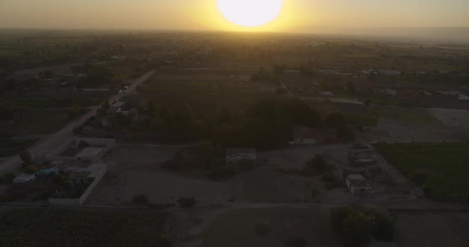 Aerial drone flies over a rural village landscape with farms and houses towards a bright hazy sunset