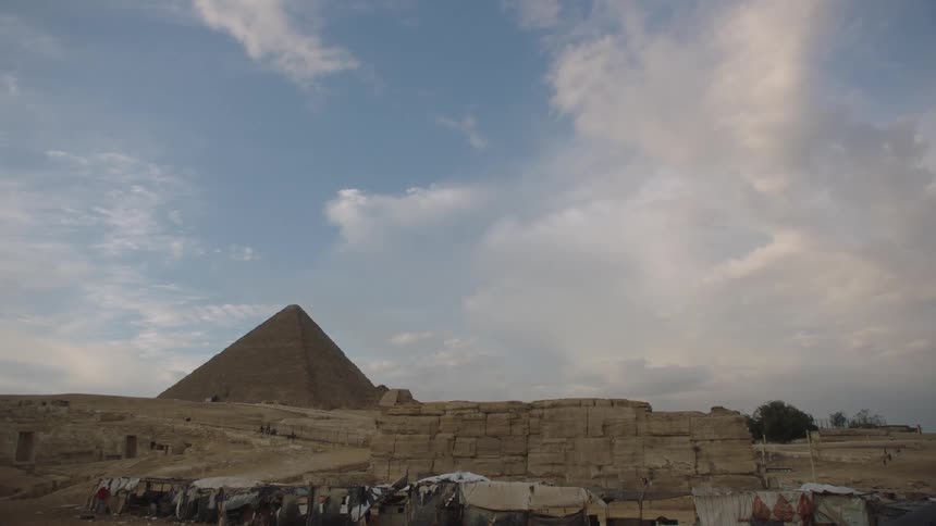 An ancient Great Pyramid of Giza looms over desert ruins and modern stalls under a vast, cloudy sky
