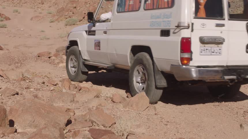 A white off-road tour vehicle navigates a rugged and rocky desert path in slow motion
