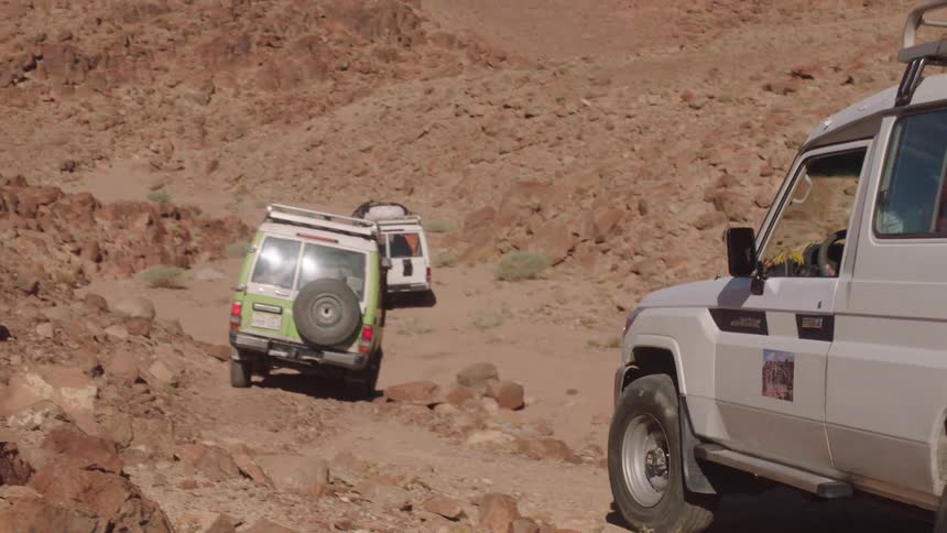 A convoy of white safari tour SUVs carefully navigates a winding, rocky road through a vast desert canyon