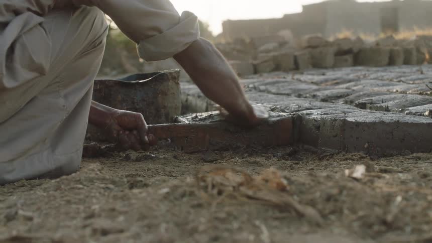 A laborer carefully shapes wet mud into a brick using a traditional wooden mold in a sunlit, dusty outdoor setting