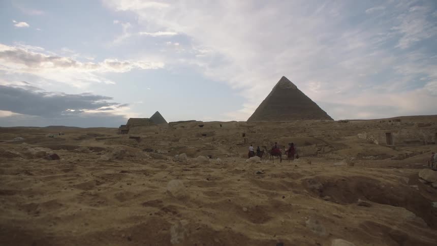 A wide shot of tourists riding camels in the vast desert with the Great Pyramids of Giza in the background