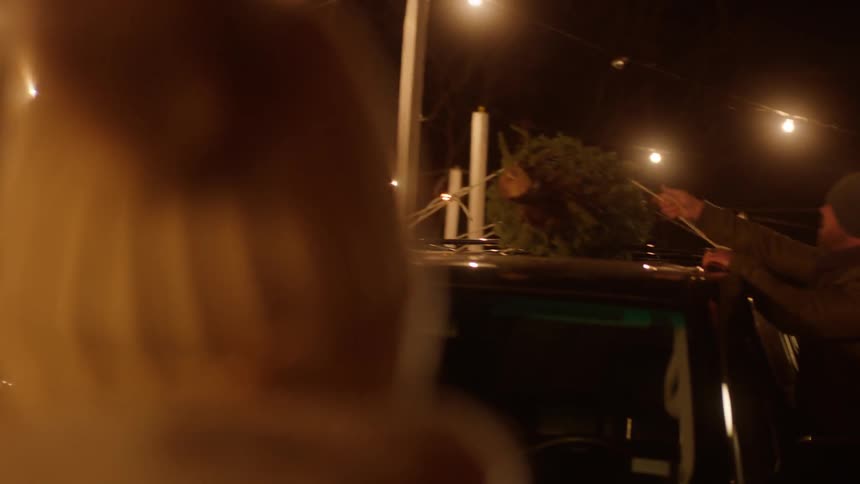 A man ties a tree to the top of a car at night