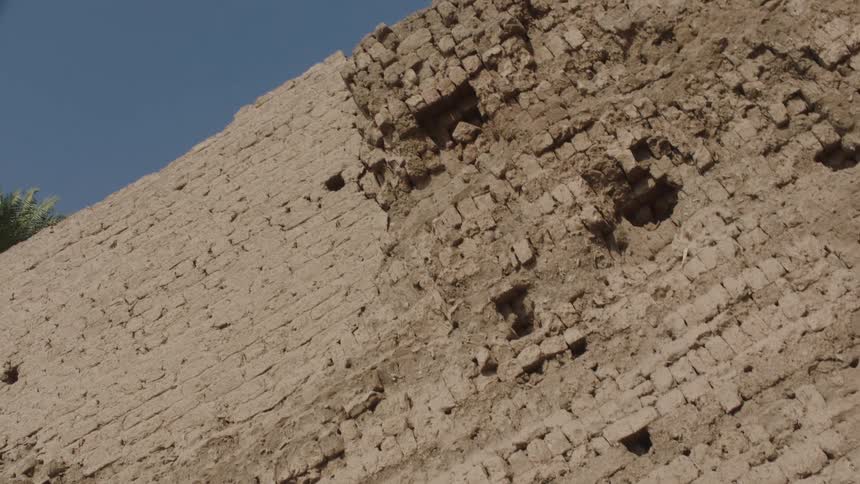 A close-up shot slowly pulls back from a crumbling ancient mud-brick wall, revealing a clear blue sky