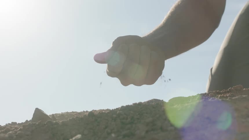 A dramatic slow-motion close-up of a hand sifting dry soil through its fingers against a bright, sun-flared sky