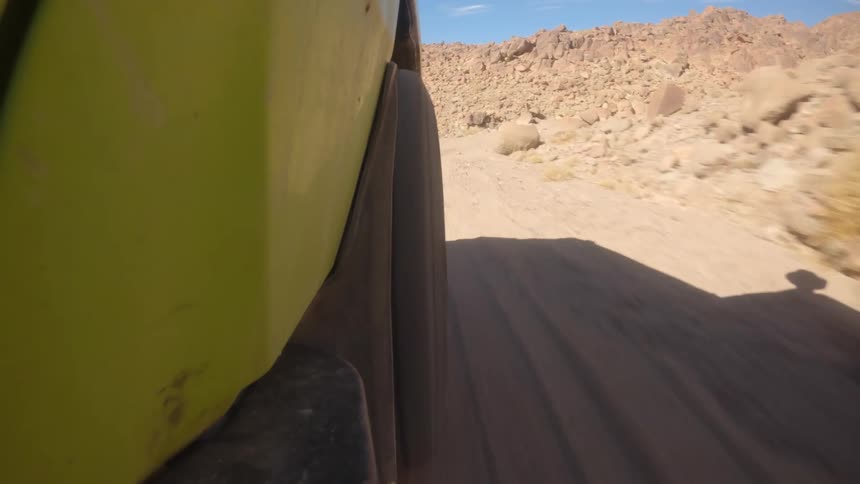 A point-of-view shot from a yellow off-road vehicle shows its tire spinning along a sandy desert road surrounded by rocky mountains