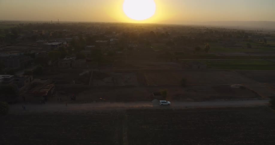 A white SUV kicks up dust as it drives along a rural dirt road through farmlands during a beautiful golden sunset