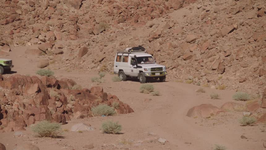 A convoy of 4x4 safari trucks carefully navigates a winding dirt road through a rugged and rocky desert canyon