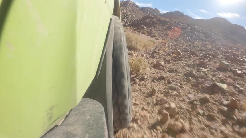 A low-angle, close-up shot of an off-road vehicle's tire as it navigates a rugged, sandy, and rocky desert trail on a sunny day