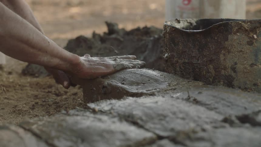 A close-up slow-motion shot shows a craftsman's hands carefully molding wet clay and straw into an adobe brick using a traditional form