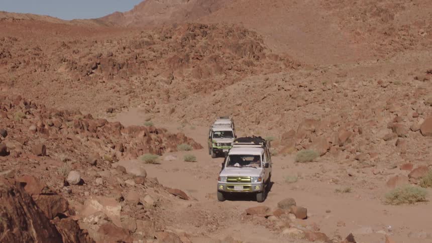 A convoy of white 4x4 off-road vehicles drives on a dirt track through a vast, rocky desert landscape