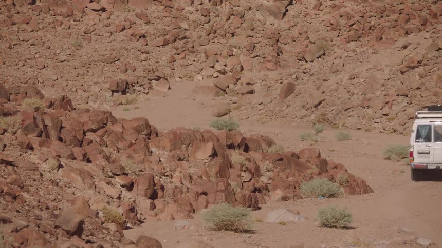 A convoy of off-road safari vehicles drives along a dusty, winding road through a rugged and rocky desert canyon