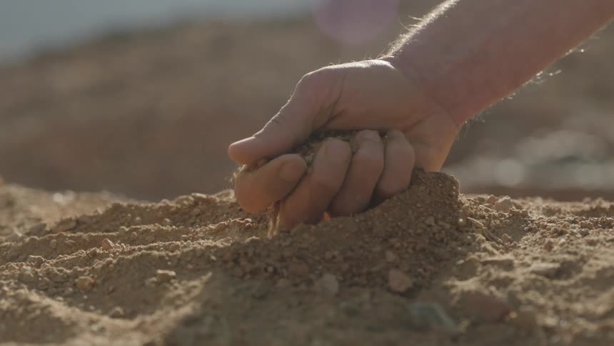 A man's hand scoops up a handful of dry soil and lets the dust and grains sift through his fingers in slow motion