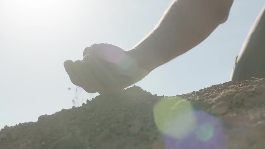 A silhouetted hand scoops up dry soil and lets it sift through their fingers in slow motion against a bright sun