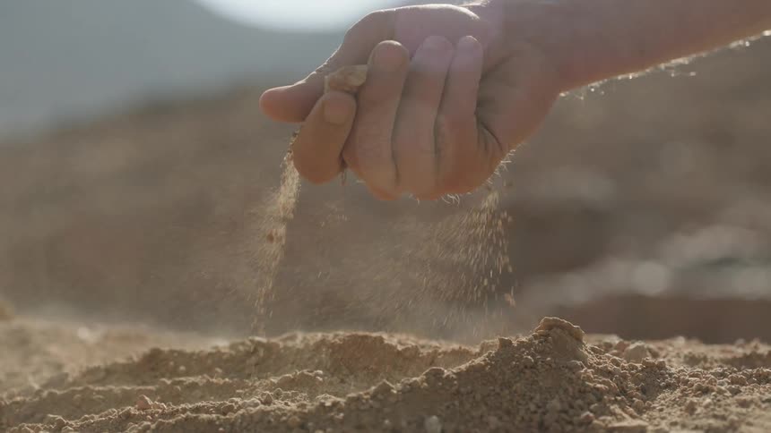 A cinematic slow-motion close-up of a person's hand letting dry, dusty earth fall through their fingers