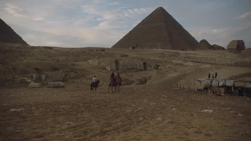 People ride camels and a horse through the Giza desert, with the Great Pyramids and Sphinx in the background