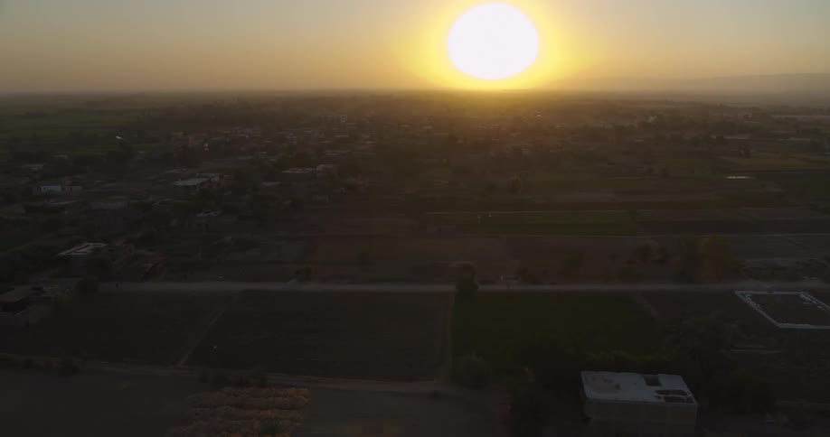 An expansive aerial shot slowly flies over a rural farming village as the sun glows brightly on the horizon at dusk