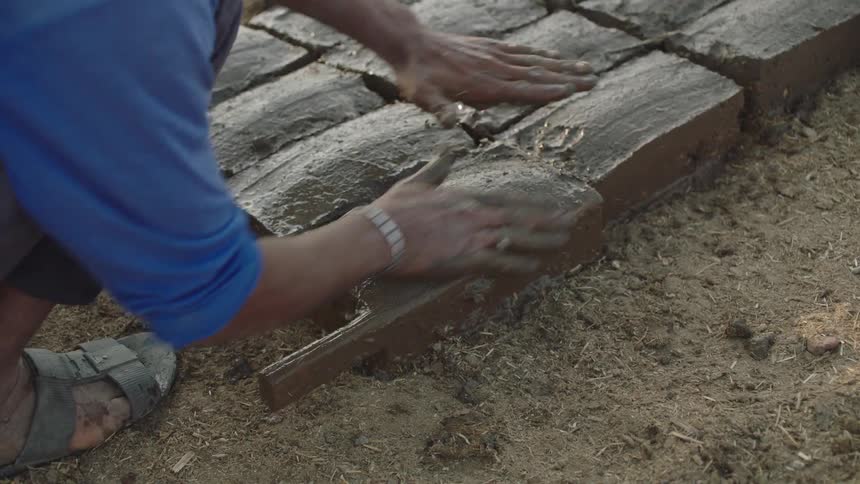 A worker's hands expertly mold and smooth wet mud and straw into adobe bricks using a traditional wooden form in slow motion