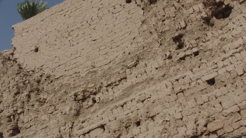 Slow tilt shot revealing the weathered texture of an ancient mud-brick wall under a clear blue sky