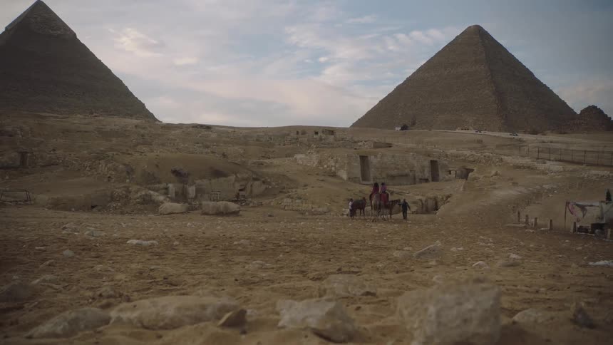 A wide, low-angle view shows locals with donkeys standing amongst ruins in the desert with the Great Pyramids of Giza in the background