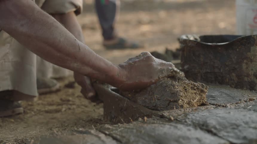 In slow motion, a laborer meticulously mixes mud and straw with their hands before pressing it into a mold to create a traditional mud brick