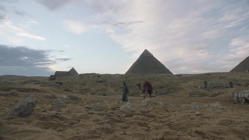 An Egyptian man in traditional clothing walks through the desert with his camel and the Great Pyramids of Giza in the background