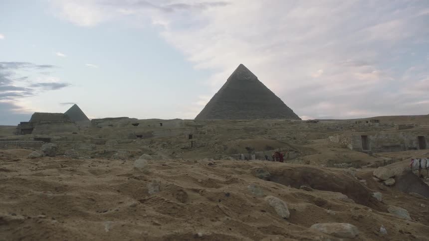A low-angle dolly shot moves across the sandy Egyptian desert towards the Great Pyramids of Giza at dusk