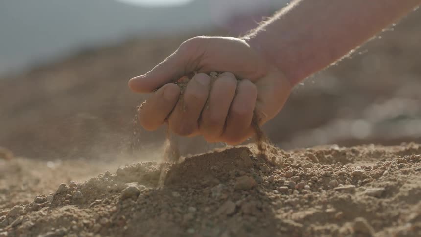 A sunlit hand scoops up a handful of dry earth and lets the dust fall through its fingers in super slow motion