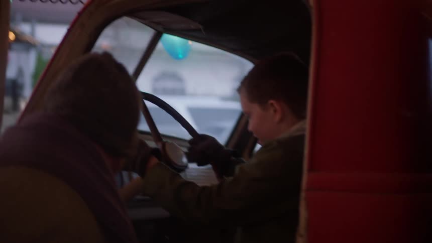 A boy climbs into the driver's seat of a truck while a man watches