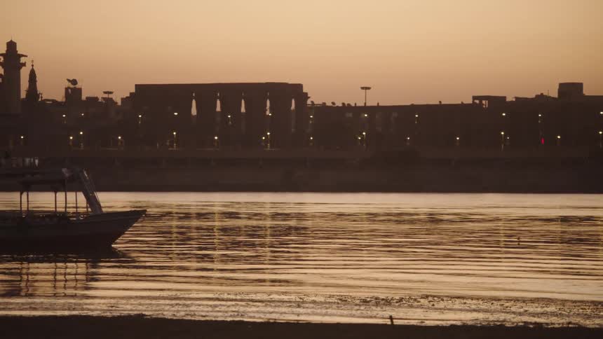 A silhouetted tour boat glides along the calm Nile River at sunset with the ancient Luxor Temple illuminated in the background