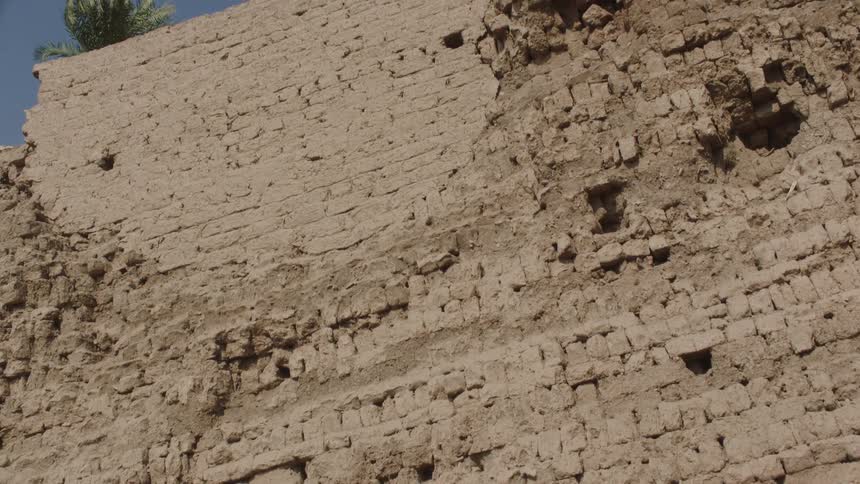 A slow upward tilt reveals the weathered texture of an ancient, crumbling mud brick wall under a clear blue sky