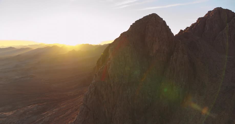 A serene static shot shows the sun rising over a vast, rocky mountain range in the desert, creating a beautiful lens flare