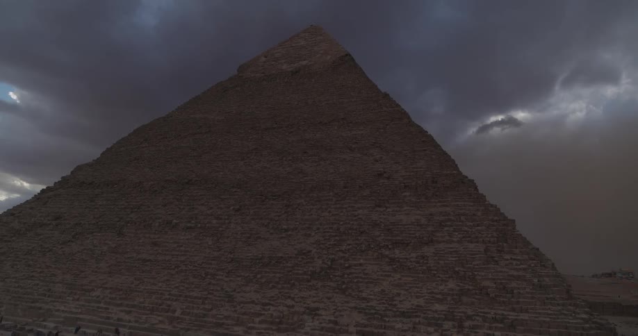 A dramatic zoom out reveals a crowd of tourists at the base of the Great Pyramid of Giza under a dark and stormy sky
