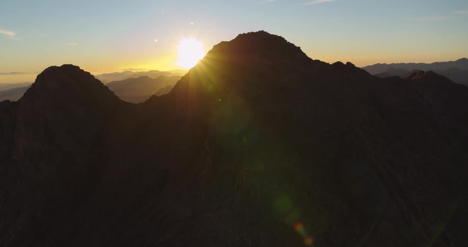 The sun slowly sets behind a dark, rugged mountain silhouette, casting a golden glow over the distant mountain range