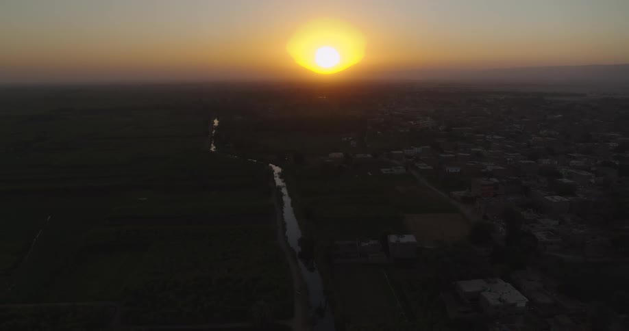 A slow pull-back aerial shot shows the golden sun setting over a river that divides a rural town and lush farmland